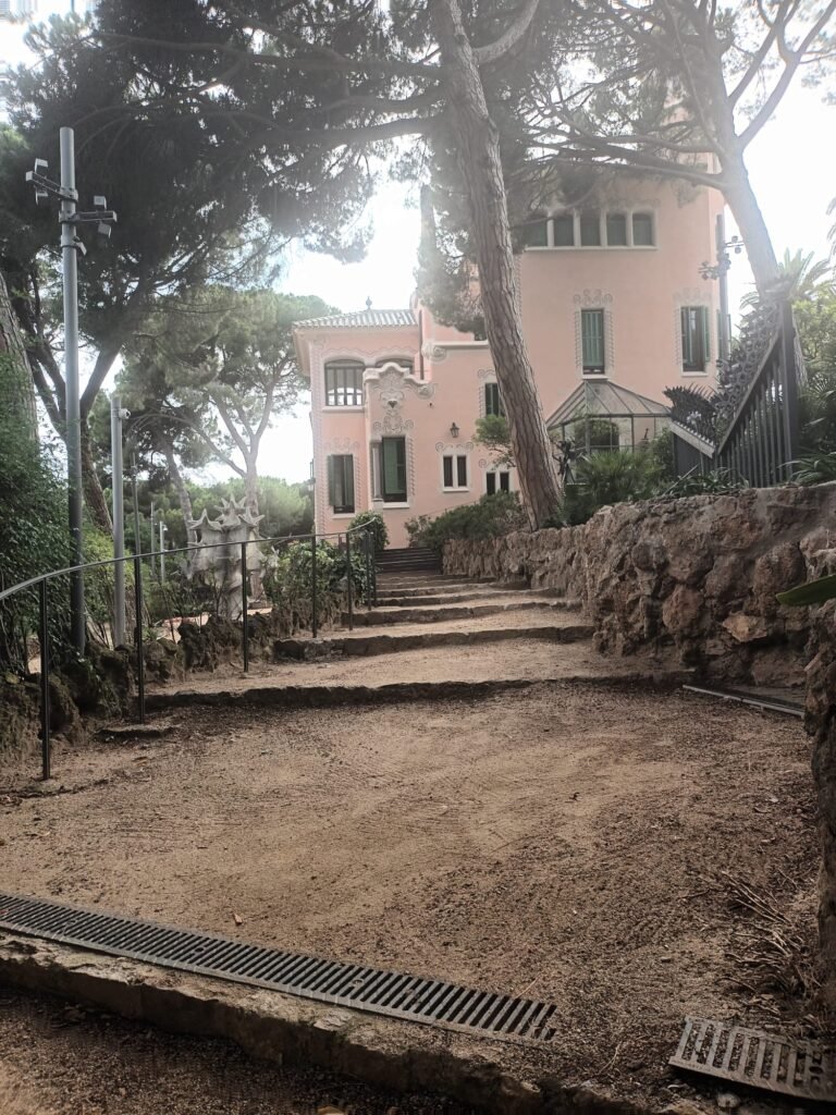 Vista de la Casa de Gaudí desde la entrada principal, rodeada de árboles y pintada de rosa