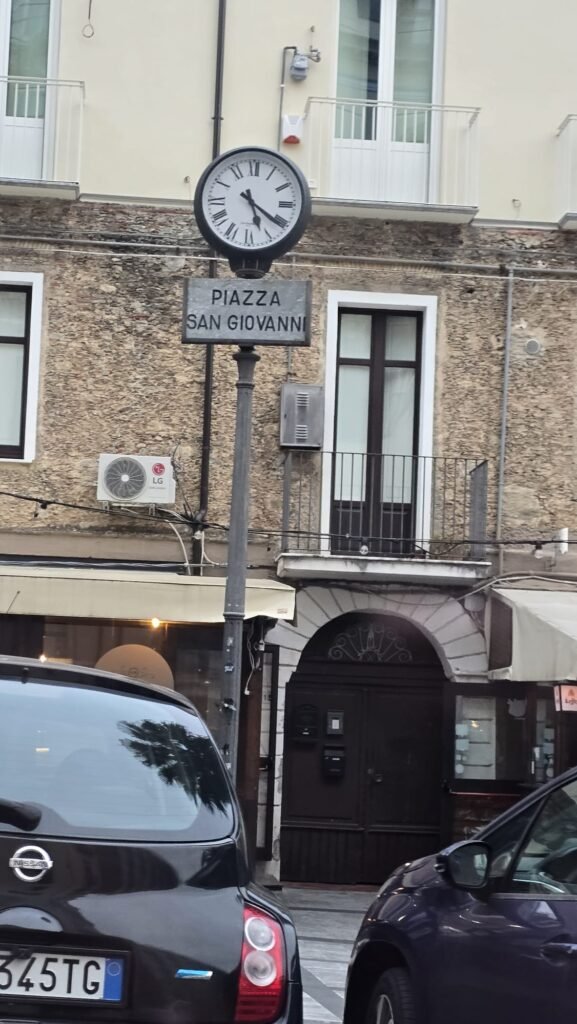 "Reloj antiguo sobre una farola junto al cartel que indica Piazza San Giovanni en Lamezia Terme, Italia"