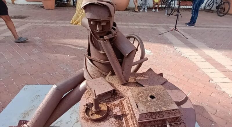 Escultura de un hombre de hojalata sentado en una silla de hierro, frente a una mesa con libros, una vela y otros objetos metálicos, en una plaza al aire libre.