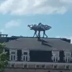 Escultura metálica de figuras humanas cargando una forma abstracta brillante, ubicada sobre el techo de un edificio clásico bajo un cielo azul con nubes.