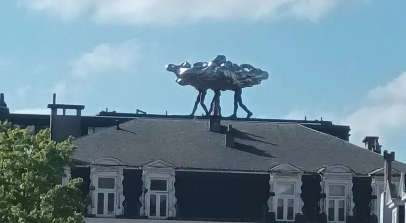 Escultura metálica de figuras humanas cargando una forma abstracta brillante, ubicada sobre el techo de un edificio clásico bajo un cielo azul con nubes.